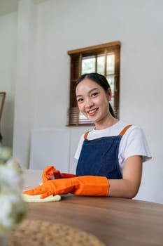 Asian woman wear rubber protective orange gloves sit clean guest reception desk with product and equipment cleaner at home.