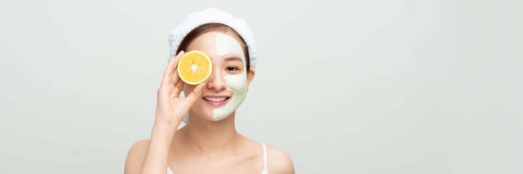 Young woman with facial clay mask holding orange fruit slice covering eyes. Banner