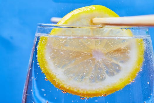 Image of Close shot of a lemon slice on a stick dangling in a fizzy glass of bubbles with a blue background