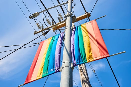 Image of Upward view of two rainbow banners on metal street light and telephone nearby with wires