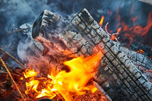 Image of Close up of smoky pit with bright yellow flames consuming leaves and three ashen white logs