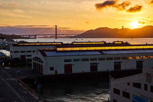 Image of Sun setting over mountains behind Golden Gate Bridge with view from warehouses on pier