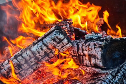 Image of Orange and yellow flames surrounding three charred black logs with white ashes in circular pit