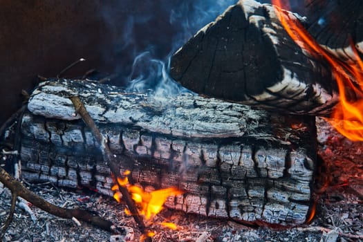 Image of Burnt logs with orange and yellow flames and smoke rising up close up background asset