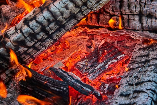 Image of Close up of red embers beneath burning and charred black logs with white ashes and tiny flames