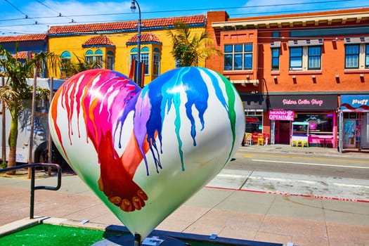 Image of Storefronts behind giant white heart LGBTQ+ sculpture with rainbow colors and hand holding