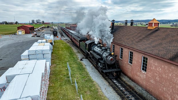 A vintage steam locomotive emits steam while departing from a brick station. Nearby, warehouses and freshly stacked materials are visible, creating a historic railroad atmosphere.
