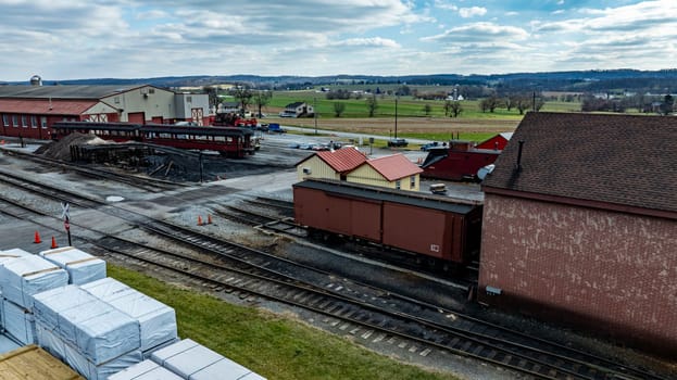 The view captures a train yard featuring several cargo cars parked near storage buildings. The setting shows a mix of industrial activity against a backdrop of green fields and a cloudy sky.