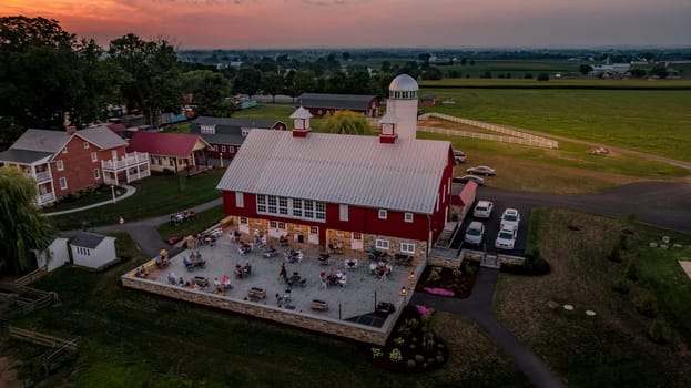 Ronks, Pennsylvania, USA, August 1, 2024 - A vibrant red barn features outdoor seating where people gather, with fields stretching into the sunset background.