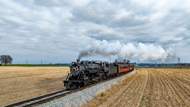 A vintage steam train chugs along railway tracks, releasing plumes of vapor as it passes through golden fields of harvested crops under a gray, overcast sky.