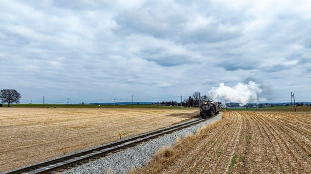 A historic steam train chugs along the tracks, billowing white smoke as it cuts through open fields under a dramatic sky filled with clouds.