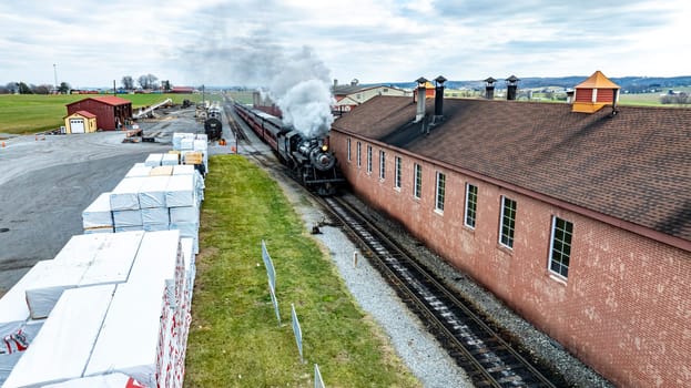 A vintage steam train puffs white smoke as it moves along the tracks next to a red brick building in a quaint rural area under cloudy skies.