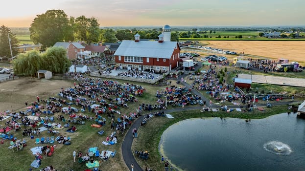 Ronks, Pennsylvania, USA, July 3, 2024 - Aerial View Of A Farm Event Featuring A Red Barn, Outdoor Orchestra, Large Crowd, Food Trucks, Parked Cars, And Surrounding Fields.