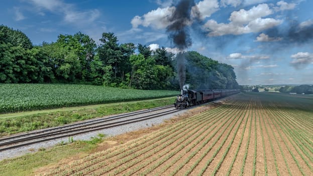 A Black Steam Locomotive Billowing Thick Smoke Pulls Red Passenger Cars Through Lush Green Fields And Forested Areas, Under A Clear Blue Sky With Puffy Clouds, Creating A Picturesque Countryside Journey.