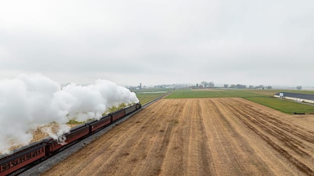 A vintage steam train chugs along tracks cutting through golden fields under a gray sky, with farmland visible in the distance, showcasing a serene rural landscape.