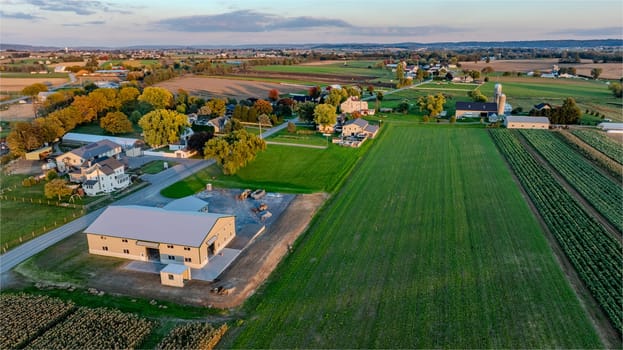 A serene rural landscape showcases green fields, a mix of houses, barns, and farms under a colorful evening sky. The tranquil setting captures the charm of countryside living.