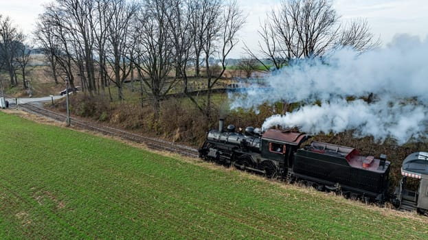 Steam Train Chugging Through a Green Field in Early Spring Near Tranquil Rural Landscape. High quality photoA classic steam train is moving along tracks bordered by lush green fields and bare trees. The smoke billows into the clear sky as it journeys through the serene countryside.