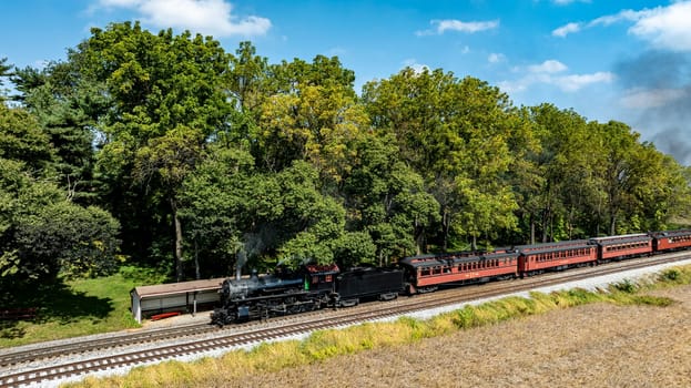 A vintage steam locomotive pulls a charming passenger train along the tracks, surrounded by vibrant trees and a clear blue sky. It creates a nostalgic atmosphere on this beautiful day.