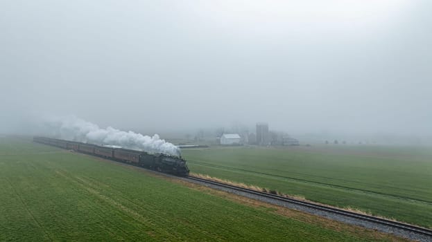 A steam locomotive moves along the tracks, shrouded by thick fog, with fields and farm structures partially visible in the background.
