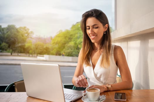 Smiling young female freelancer stirring coffee in cup while looking at laptop screen. Beautiful businesswoman sitting in an online meeting while having drink at outdoor cafe.