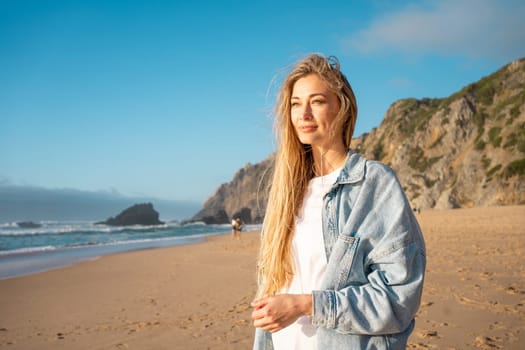 Blond woman in denim shirt looking away while standing on sandy beach. Thoughtful lady with long hair near sea and mountain in background enjoying summer vacation.