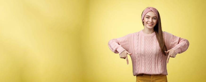 Tender young 20s european girl in sweater smiling broadly finally take-off braces pointing down promoting skillful dentist professional. Female student standing happy positive over yellow wall.