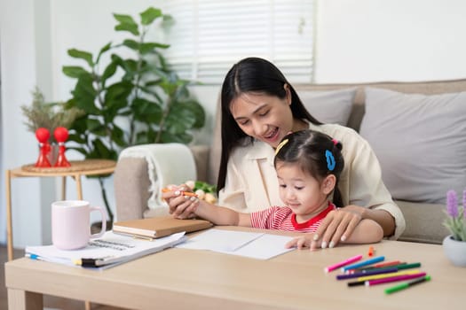 Asian mother lovingly guides her young daughter in a drawing activity, fostering creativity and bonding in a warm home setting.