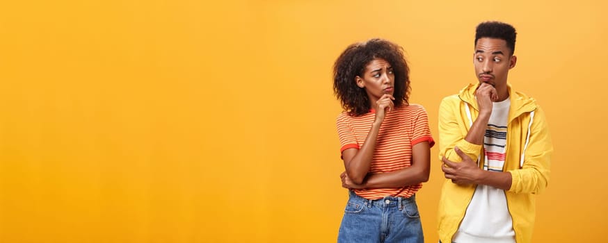 Silly confused girl standing gloomy and worried pursing lips and frowning holding hand on chin looking at boyfriend confused while guy glancing at girlfriend with curious expression over orange wall. Lifestyle.