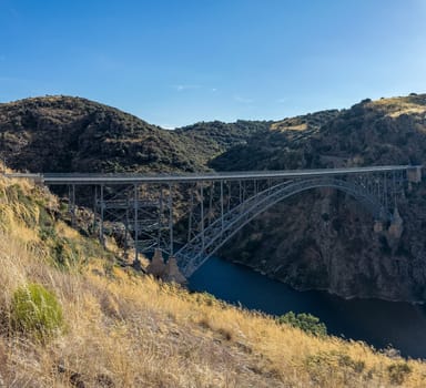 A steel arch bridge crosses a tranquil river, surrounded by rugged hills beneath a clear sky.