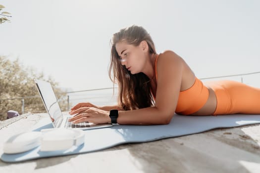 A woman wearing an orange sports bra and leggings lies on a yoga mat while working on a laptop in a sunny outdoor setting.