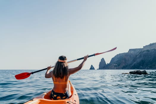 A woman paddles a kayak towards a rocky cliff in the ocean. The sun shines brightly and the water is calm.