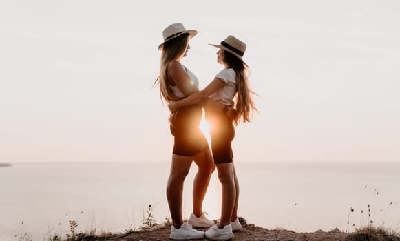 Two women in hats stand on a cliff overlooking the ocean, embracing as the sun sets behind them.
