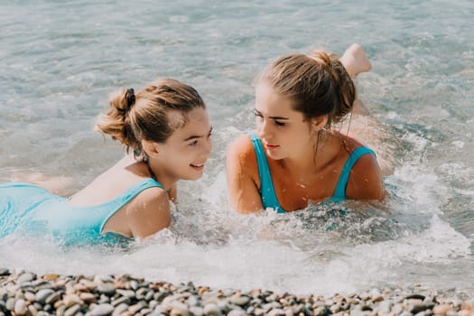 Two young women in blue swimsuits are lying in the shallows of the ocean, one looking at the camera and the other looking at her companion, their hair is wet and they are covered in water.