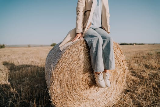 A woman wearing a trench coat and blue jeans sits on a large hay bale in a field. The sky is blue and the field is brown.