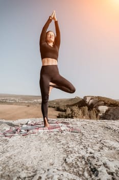 A woman in black leggings and a long-sleeved top performs a yoga pose on a rocky clifftop, with a checkered blanket beneath her.