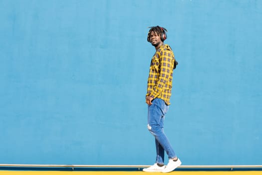attractive young black man walks around listening to music with large headphones on a blue background, concept of technology and lifestyle, copy space for text