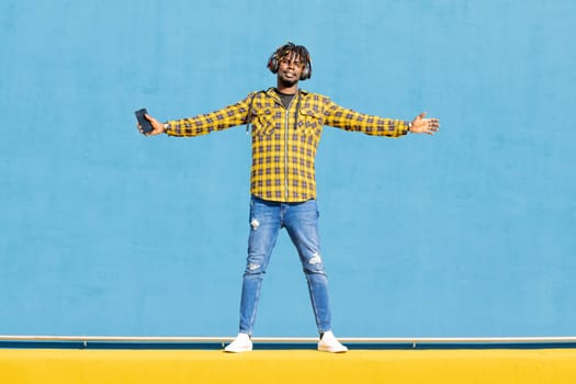 attractive young black man listening to music from his phone with a large headphone on a blue background, technology and lifestyle concept, copy space for text