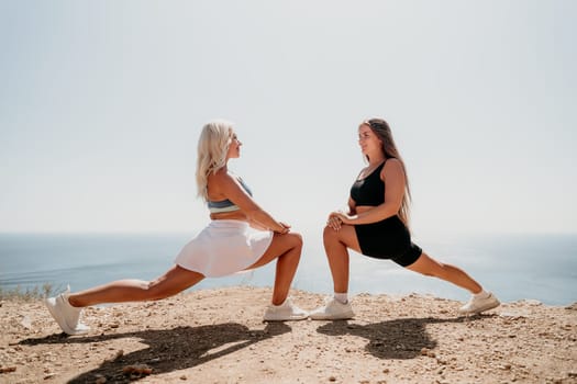 Two women in sportswear perform a forward lunge stretch on a cliffside overlooking the ocean.