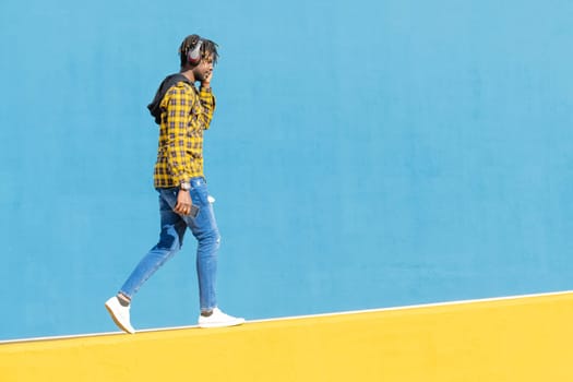 young black man with headphones walking along a yellow wall and a blue wall in the background with his smartphone in his hand, technology and lifestyle concept, copy space for text