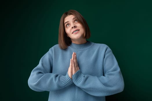 A woman wearing a blue sweater is shown in a prayerful pose, with her hands clasped together and eyes closed. She appears to be deep in prayer, with a solemn expression on her face.