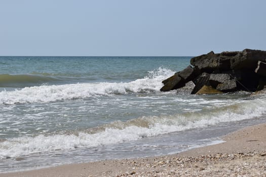 The waves of the turquoise sea break on the coastal boulders and a dense white foam is formed. Clear blue sky. White waves breaking on sandy beach through gap in rocks on sea