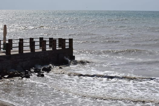 big sea waves splashing against a pier. The pier cuts the sea waves off the coast during a stormy sunny day.