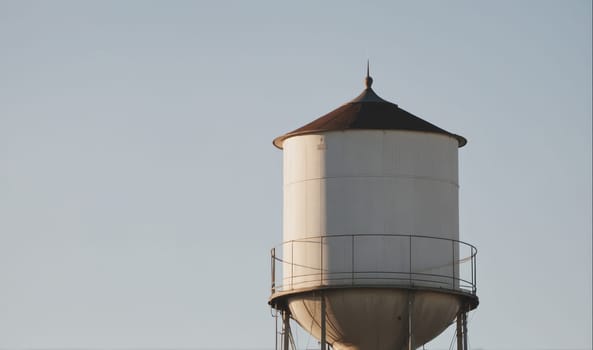 The image features a water tower with a white, cylindrical body and a black, conical roof. The tower is surrounded by a metal railing. The sky is clear and blue, with no clouds visible. The tower is positioned in the center of the frame, with the sky filling the rest of the composition.