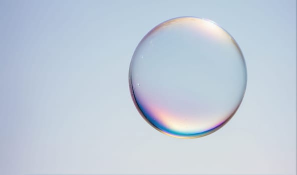 A single soap bubble floats in the air. The bubble is perfectly round and iridescent, reflecting light in a rainbow of colors. The background is a soft, light blue, which creates a striking contrast with the bubble.