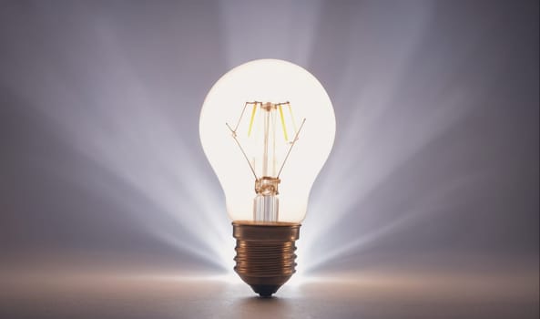 A single, clear light bulb with a copper base is lit and sitting on a beige surface. Light shines from the bulb and spreads out in the background, illuminating the dark, gray space.