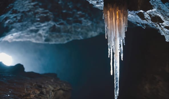 A single, long icicle hangs from the ceiling of a dark cave, illuminated by a faint light source from outside. The icicle is translucent and has a slight orange tint. The cave walls are rough and textured, with a sense of mystery and depth.