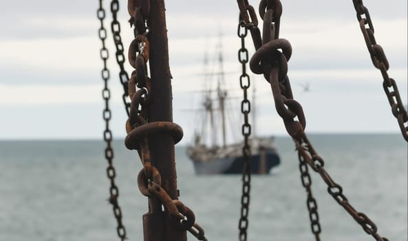 A chain link fence hangs on a boat and a large ship is in the distance. The sea is choppy. The sky is overcast.