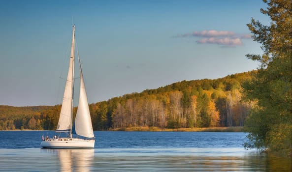 A white sailboat sails across a calm blue lake on a sunny day. The lake is surrounded by a wooded shoreline with fall foliage, and a small cloud floats in the blue sky.