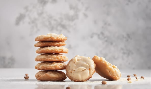 A stack of cookies is on a white countertop. The cookies are round and have a light brown color. They are stacked on top of each other in a neat pile. There is a single cookie lying to the right of the stack, and a partially eaten cookie is lying to the left of the stack. There are a few scattered almonds in front of the cookies.