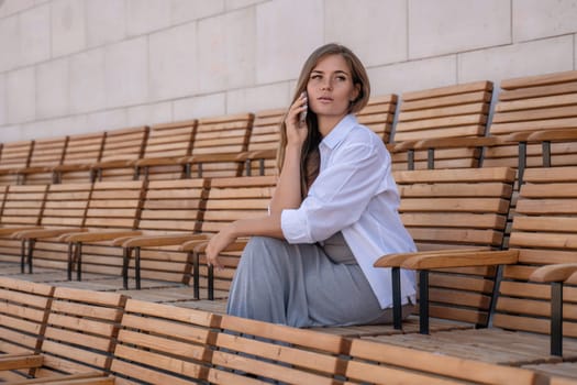 A woman in a white shirt and grey skirt sits on wooden bleachers, talking on a cell phone.